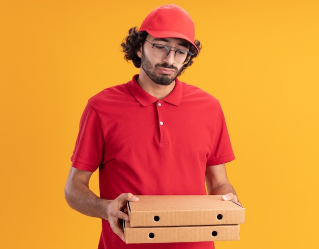 Gratis foto verwarde jonge blanke bezorger in rood uniform en pet met een bril die pizzapakketten vasthoudt en bekijkt