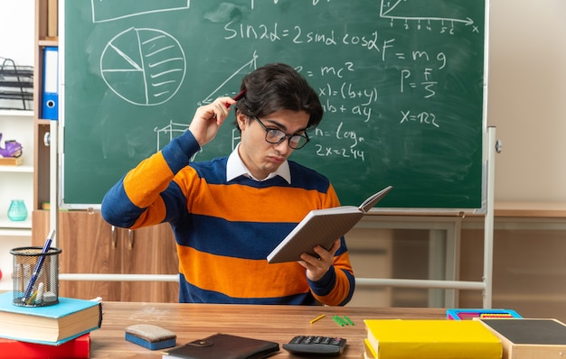 Verwarde jonge blanke geometrieleraar met een bril die aan het bureau zit met schoolhulpmiddelen in de klas die vasthoudt en naar een notitieblok kijkt die het hoofd aanraakt met een pen