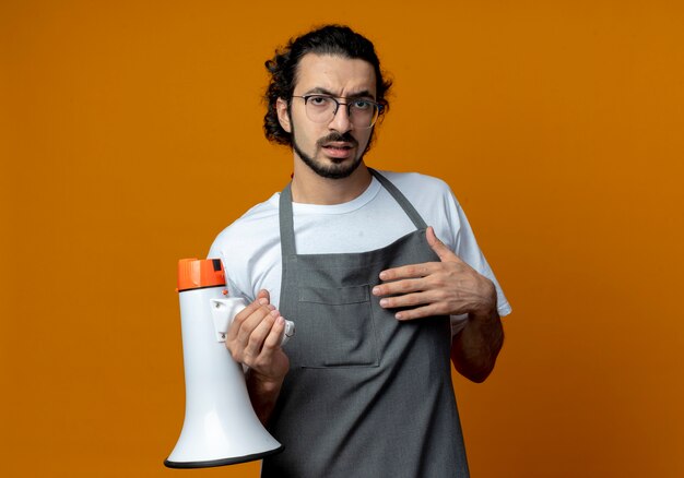 Verwarde jonge blanke mannelijke kapper dragen van uniform en bril met spreker en aanraken van borst geïsoleerd op een oranje achtergrond met kopie ruimte