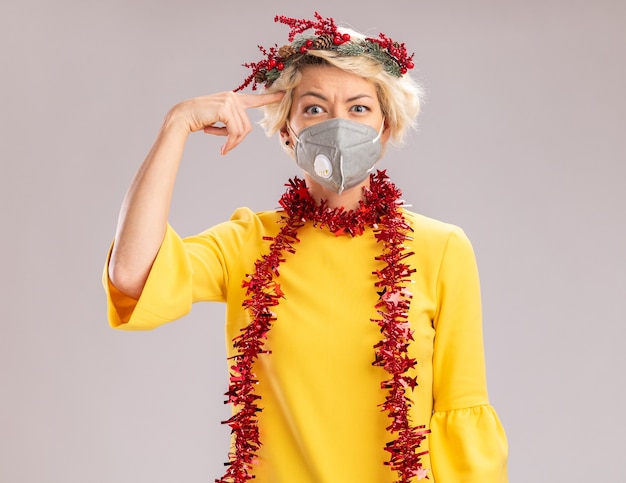 Verwarde jonge blonde vrouw die de hoofdkrans van Kerstmis en klatergoudslinger om hals met beschermend masker draagt ?? die doet denk gebaar kijkt dat op witte muur wordt geïsoleerd