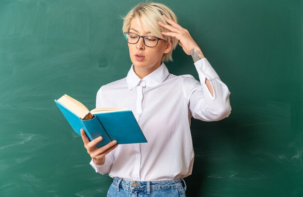 Verwarde jonge blonde vrouwelijke leraar met een bril in de klas die voor het schoolbord staat en een boek leest dat het hoofd aanraakt