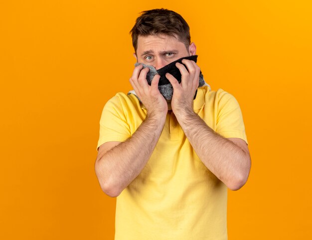 Verwarde jonge blonde zieke Slavische man die sjaal draagt en houdt die op oranje muur met exemplaarruimte wordt geïsoleerd