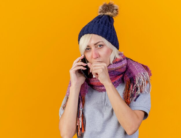 Verwarde jonge blonde zieke Slavische vrouw die de wintermuts en sjaal draagt, spreekt over telefoon die hand dicht bij mond houdt die op oranje muur met exemplaarruimte wordt geïsoleerd