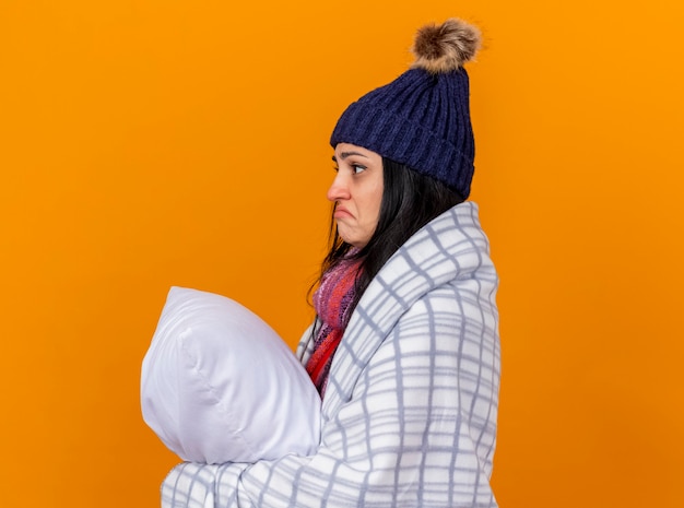 Gratis foto verwarde jonge kaukasische ziek meisje dragen winter muts en sjaal verpakt in plaid staande in profiel te bekijken met kussen op zoek recht geïsoleerd op een oranje achtergrond met kopie ruimte