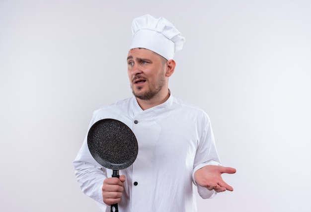 Verwarde jonge knappe kok in chef-kok uniform met koekenpan met lege hand en kijkend naar kant geïsoleerd op een witte muur white