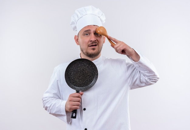 Verwarde jonge knappe kok in uniform van de chef-kok die een koekenpan vasthoudt en een lepel op het voorhoofd zet, geïsoleerd op een witte muur