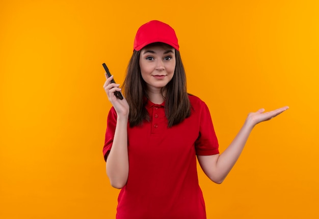 Verwarde jonge leveringsvrouw die rode t-shirt in rode pet draagt die een telefoon op geïsoleerde oranje muur houdt