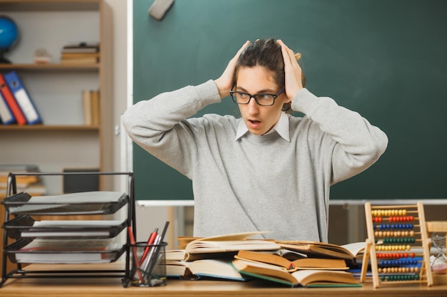 Verwarde jonge mannelijke leraar greep het hoofd terwijl hij aan het bureau zat met schoolgereedschap aan in de klas