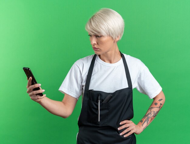 Verwarde jonge mooie vrouwelijke kapper in uniform houden en kijken naar telefoon hand op heup geïsoleerd op groene muur