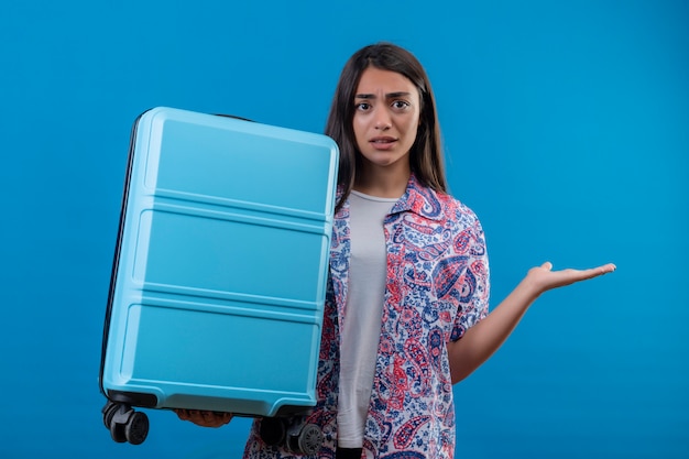 Verwarde jonge reiziger vrouw met reiskoffer twijfelachtig schouderophalend staande op geïsoleerde blauw