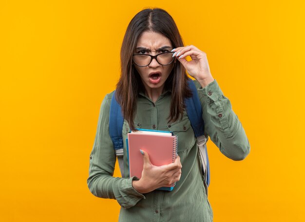 Verwarde jonge schoolvrouw die een rugzak draagt met een bril die boeken vasthoudt