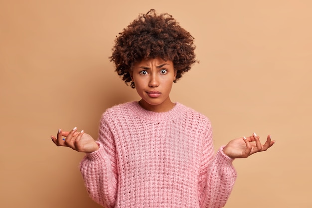 Gratis foto verwarde jonge vrouw met afro-kapsel spreidt handpalmen en gezichten dilemma heeft moeilijke keuze gekleed in casual gebreide trui geïsoleerd over bruine muur