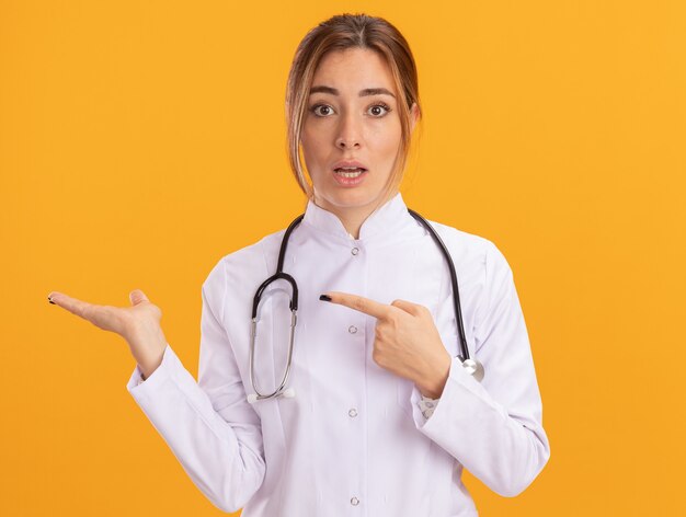 Verwarde jonge vrouwelijke arts die een medisch gewaad draagt met een stethoscoop die doet alsof hij vasthoudt en wijst naar iets dat op een gele muur is geïsoleerd
