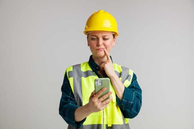 Verwarde jonge vrouwelijke bouwvakker die veiligheidshelm en veiligheidsvest draagt ?? die en mobiele telefoon bekijkt die vinger op mondhoek houdt