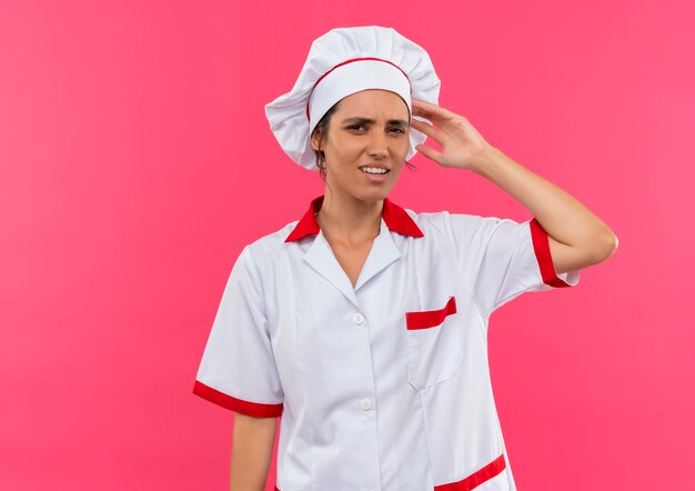 Verwarde jonge vrouwelijke kok die eenvormige chef-kok draagt die hand op hoofd op geïsoleerde roze muur met exemplaarruimte zet