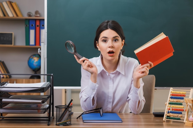 verwarde jonge vrouwelijke leraar die een boek vasthoudt met een vergrootglas dat aan het bureau zit met schoolhulpmiddelen in de klas