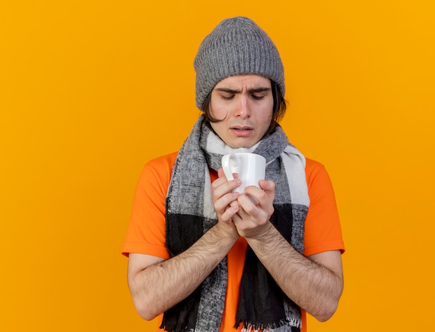 Gratis foto verwarde jonge zieke man met winter hoed met sjaal houden en kijken naar kopje thee geïsoleerd op een oranje achtergrond