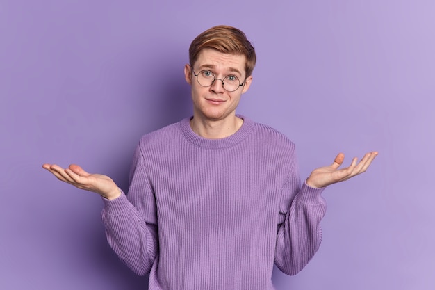 Verwarde jongere haalt zijn schouders op, spreidt zijn handpalmen, staat besluiteloos binnen, gekleed in een casual trui draagt een ronde bril