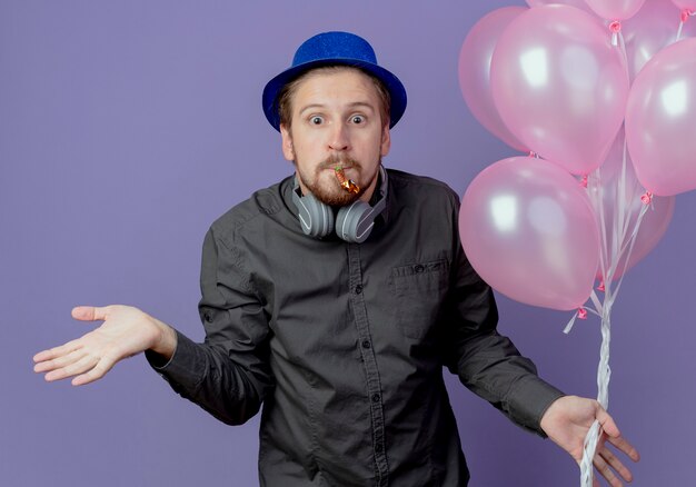 Verwarde knappe man met blauwe hoed en koptelefoon op nek staat met helium ballonnen blazen fluitje geïsoleerd op paarse muur