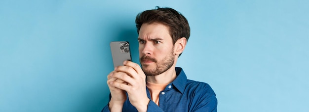 Verwarde man die dicht naar het scherm van de mobiele telefoon staart en fronsend op een blauwe achtergrond staat