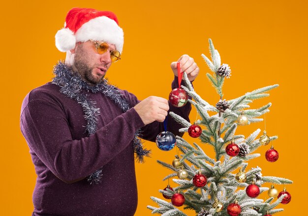 Verwarde man van middelbare leeftijd met kerstmuts en klatergoud slinger om nek met bril staande in profielweergave in de buurt van de kerstboom en versiert het met kerstballen geïsoleerd op een oranje achtergrond