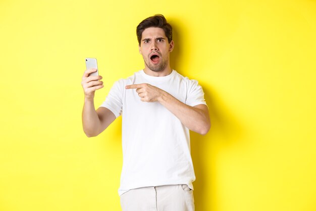 Verwarde man wijzende vinger op het scherm van de mobiele telefoon, kan iets niet begrijpen, staande op gele achtergrond. Kopieer ruimte