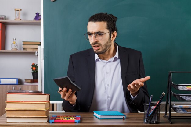 Verwarde mannelijke leraar met een bril die een rekenmachine vasthoudt en bekijkt die aan tafel zit met schoolhulpmiddelen in de klas