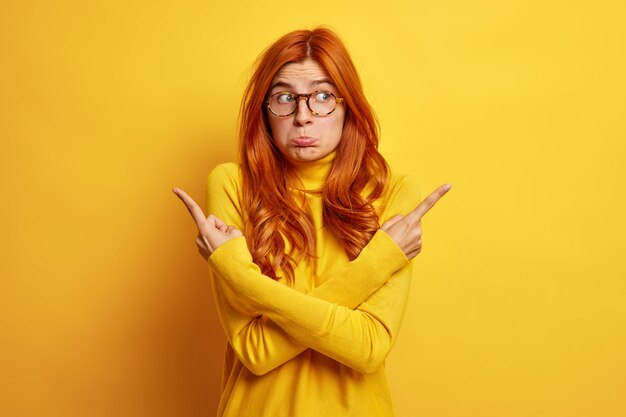 Gratis foto verwarde ongelukkige vrouw met lang rood haar kruist armen over lichaam en wijst naar verschillende kanten aarzelt wat beter te kiezen draagt gele trui