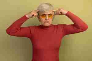 Gratis foto verwarde oude vrouw met een rode coltrui en een zonnebril die er recht uitziet en een denkgebaar doet dat op een olijfgroene muur is geïsoleerd