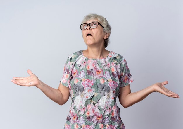 Verwarde oudere vrouw in optische bril houdt handen open opzoeken geïsoleerd op witte muur