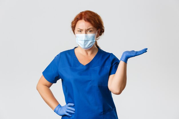 Verwarde sceptische vrouwelijke arts, tandarts in scrubs, gezichtsmasker en handschoenen, schouderophalend, naar rechts wijzend en fronsen teleurgesteld
