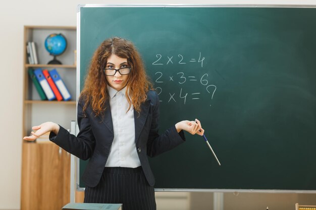 verwarde spreidende handen jonge vrouwelijke leraar met aanwijzer die vooraan op het schoolbord staat in de klas