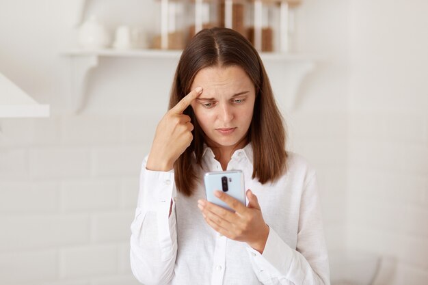 Verwarde verwarde donkerharige vrouw die mobiele telefoon in handen heeft, sociale netwerken controleert, negatief bericht of commentaar leest, thuis in de keuken poseert.