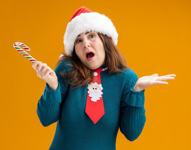 Verwarde volwassen blanke vrouw met kerstmuts en santa stropdas bedrijf candy cane geïsoleerd op een oranje achtergrond met kopie ruimte