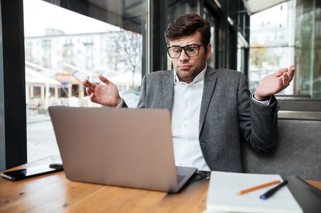 Verwarde zakenman die in oogglazen door de lijst in koffie zitten terwijl zijn schouders ophaalt en laptop computer bekijkt