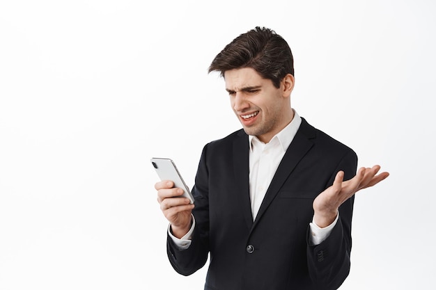 Verwarde zakenman die naar smartphone kijkt met een verbaasd gezicht, een vreemd nieuwsbericht leest, staande tegen een witte achtergrond in een zwart pak