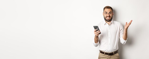Verwarde zakenman die vreemd bericht op mobiele telefoon leest en er geïrriteerd uitziet terwijl hij over een witte achtergrond staat