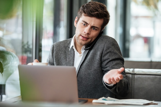 Verwarde zakenmanzitting door de lijst in koffie met laptop computer terwijl het spreken door smartphone