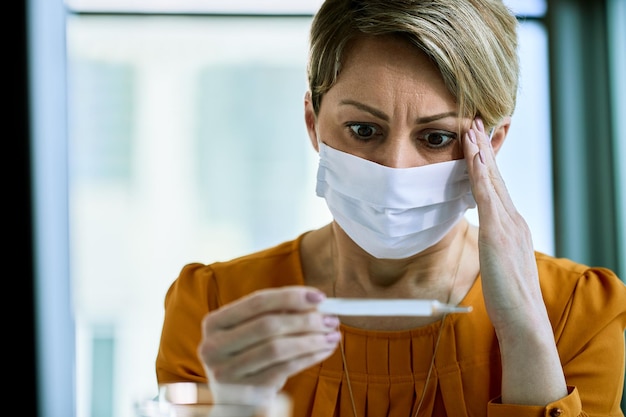 Verwarde zakenvrouw die een gezichtsmasker draagt tijdens het gebruik van een thermometer en het meten van haar temperatuur op kantoor