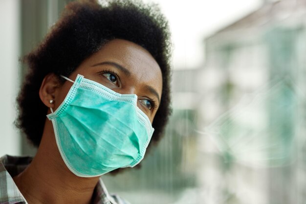 Verwarde zwarte vrouw met gezichtsmasker die denkt terwijl ze thuis door het raam kijkt