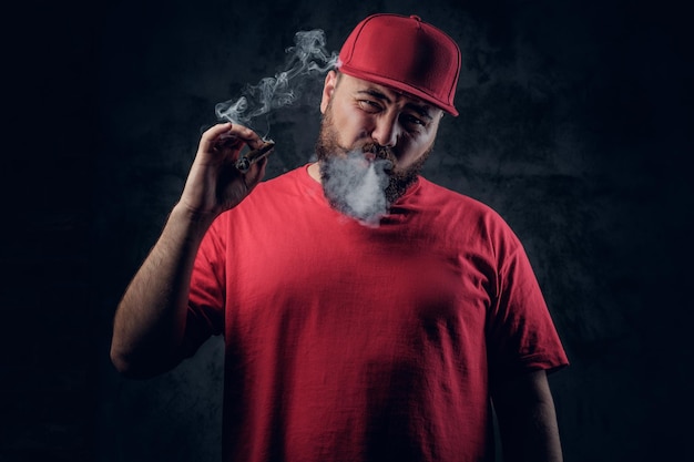 Vet bebaarde hipster man in een rode hiphop kleding roken van een sigaret op een grijze achtergrond.