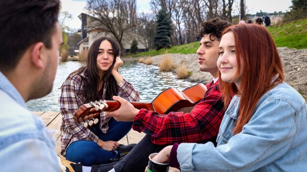 Vier jonge vrienden zingen, rusten en spelen gitaar in de buurt van een meer in een park