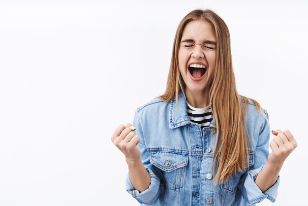 Viering prestatie en doelen concept vrolijk gelukkig blond meisje vieren overwinning schreeuw ja vreugde balde vuisten en ogen gesloten opgelucht gevoel vrolijk winnende prijs witte achtergrond