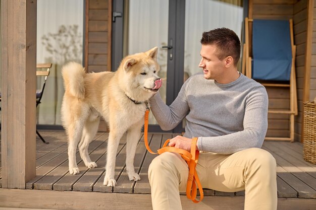 Viervoetige vriend. Jonge volwassen glimlachende man in vrijetijdskleding die op de veranda van het landhuis zit en uitkijkt terwijl hij op mooie dag de gemberhond met leiband aanraakt