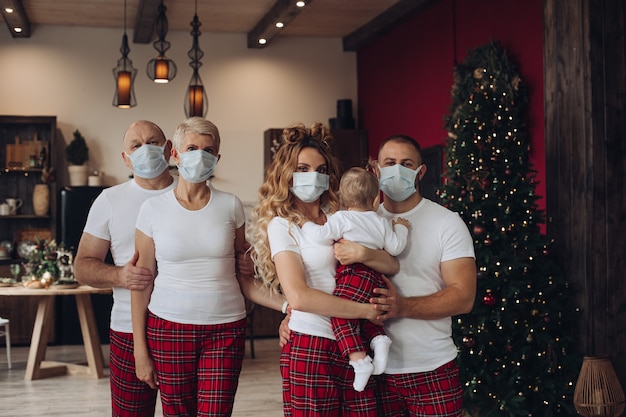 Vijf familieleden met een baby in t-shirts en broek in beschermende gezichtsmaskers met Kerstmis thuis.