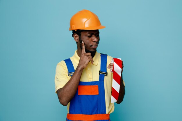 Vinger op de wang met duct tape jonge Afro-Amerikaanse bouwer in uniform geïsoleerd op blauwe achtergrond