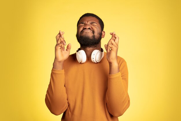 Vingers gekruist, geluk. Portret van een jonge Afro-Amerikaanse man op geel, gezichtsuitdrukking. Mooi mannelijk model met koptelefoon, copyspace. Concept van menselijke emoties, verkoop, advertentie.