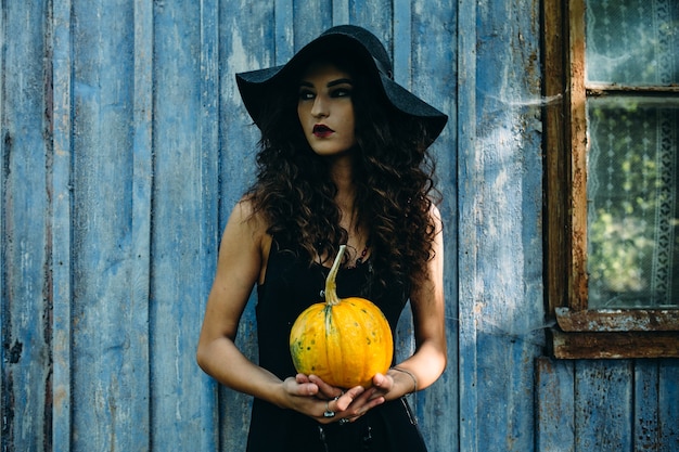 Vintage vrouw als heks poseren met pompoen in de handen tegen verlaten plek aan de vooravond van Halloween