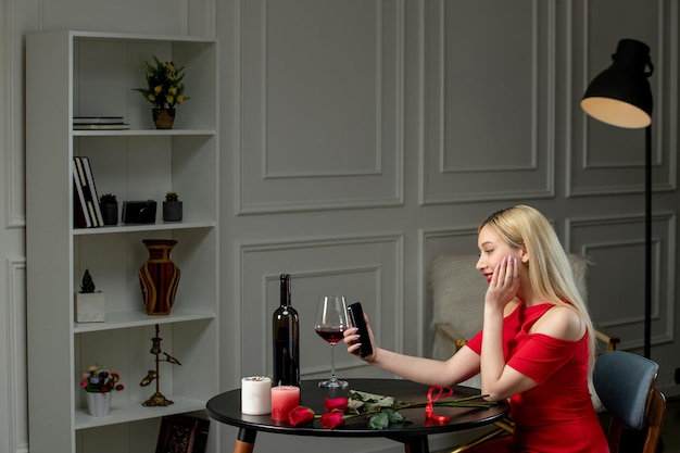 Virtueel liefdes schattig blond meisje in rode jurk op afstandsdatum met wijn en kaarsen met mobiele telefoon