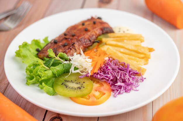 Vis steak met frietjes, kiwi, sla, wortelen, tomaten en kool in een witte schotel.